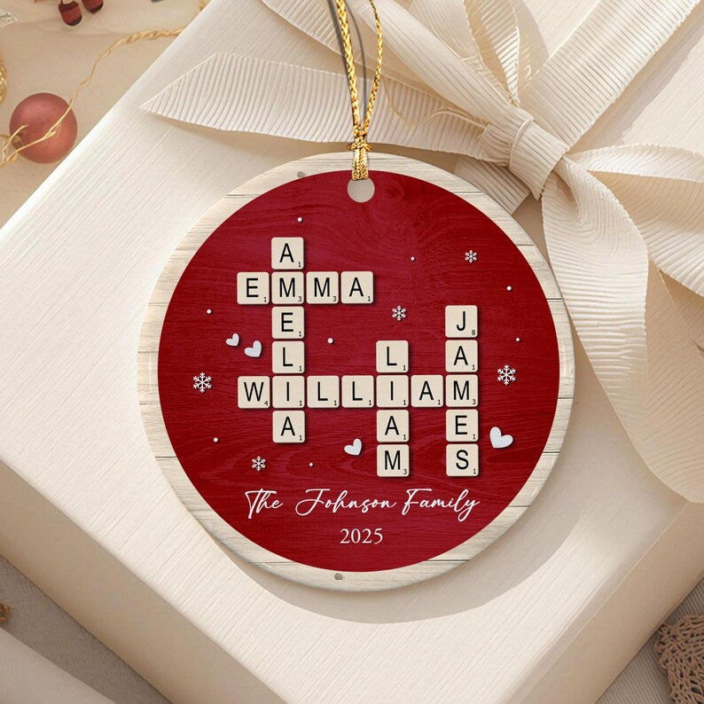 A round, red, wood-textured Christmas ornament featuring a crossword-style arrangement of letter tiles spelling AMELIA, EMMA, WILLIAM, and JAMES.
