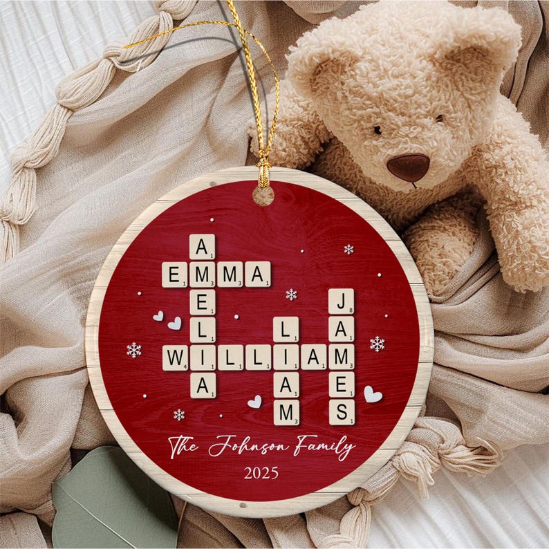 A round, red, wood-grain patterned ornament featuring a crossword-style arrangement of letter tiles spelling the names Emma, Amelia, William, Liam, and James. The text The Johnson Family and 2025 are printed below the tiles.