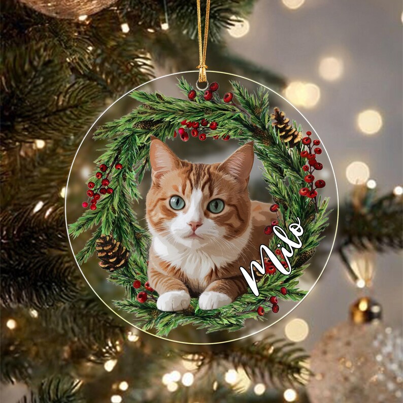 A round, flat Christmas ornament featuring a ginger and white cat centered within a wreath of green pine needles, red berries, and brown pinecones.