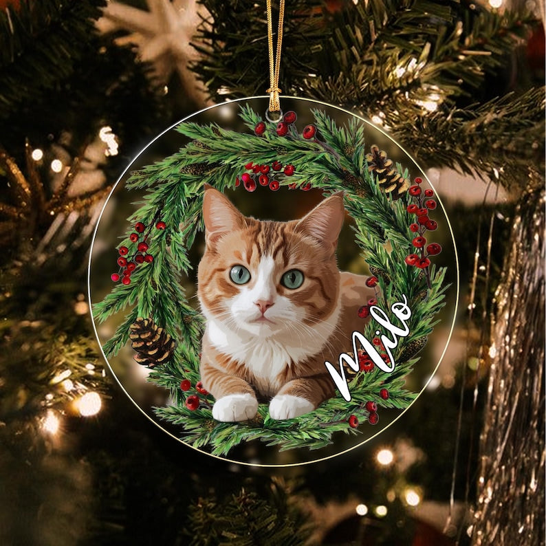 A round Christmas ornament featuring a ginger and white cat centered within a wreath of green pine needles, red berries, and brown pinecones. The name Milo is written in white cursive script on the right side of the wreath.