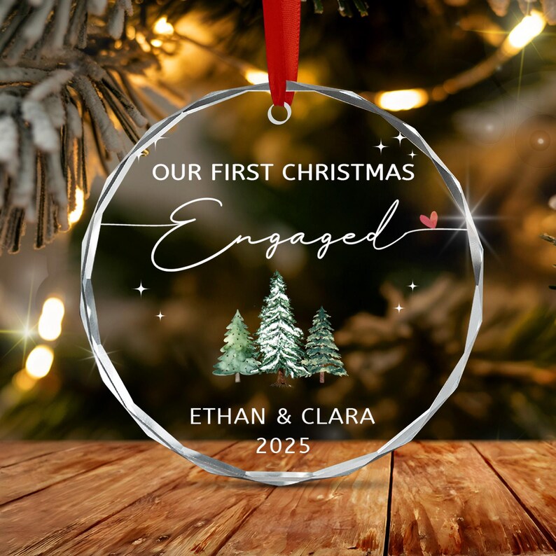 A circular clear glass ornament with faceted edges, featuring a red ribbon hanger. The text reads "OUR FIRST CHRISTMAS Engaged" with a small red heart, three green watercolor pine trees, and the names "ETHAN & CLARA" with "2025".