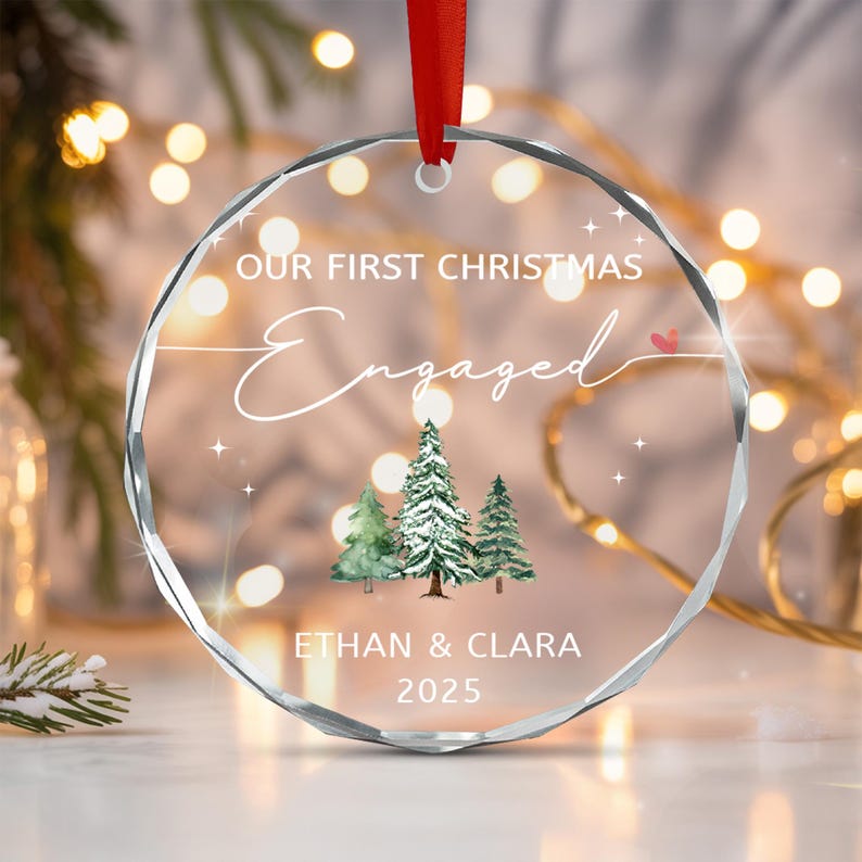 A circular clear acrylic ornament with faceted edges, featuring a red ribbon hanger. The text reads OUR FIRST CHRISTMAS Engaged, ETHAN & CLARA, and 2025.