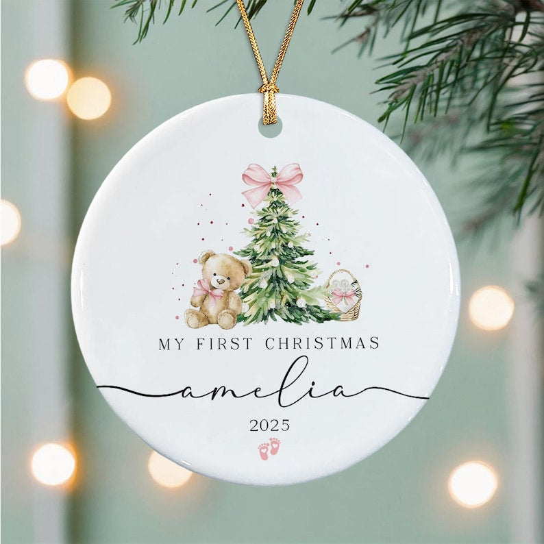 A round white ceramic ornament hanging from a gold string. The design features a watercolor illustration of a teddy bear, a Christmas tree with a pink bow, and a basket.