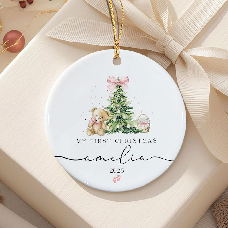 A round white ceramic ornament featuring a watercolor illustration of a green Christmas tree with a pink bow, a teddy bear, and a basket.