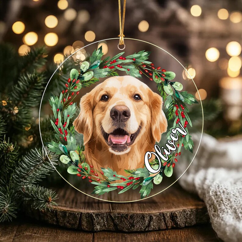 A circular clear acrylic ornament featuring a golden retriever portrait framed by a watercolor-style wreath of green leaves and red berries. The name Oliver is printed in white cursive script on the right side of the wreath.