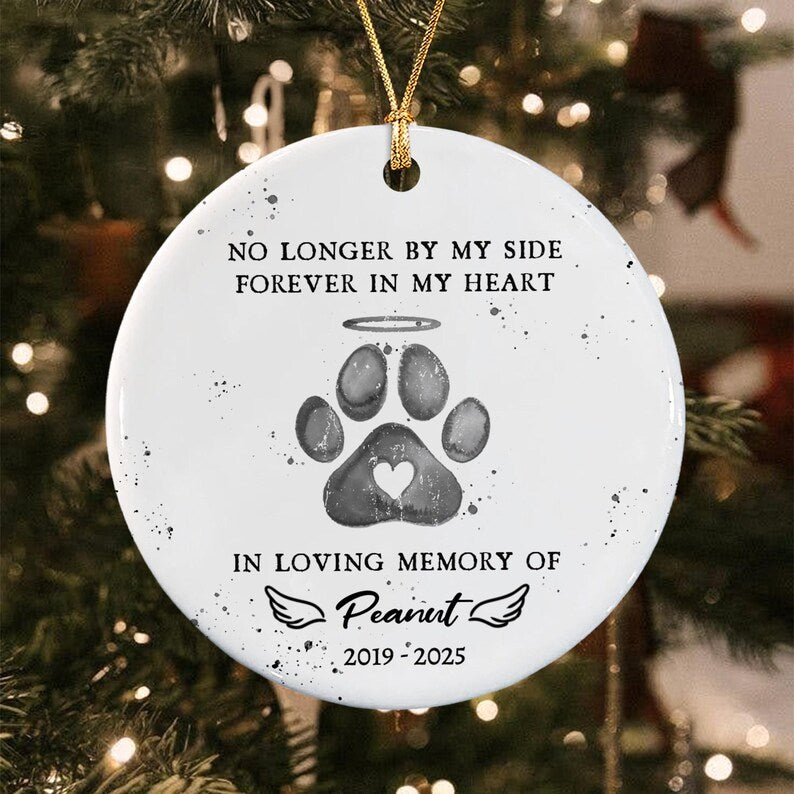 A round white ceramic ornament featuring a black paw print with a halo and a small heart cutout. The text reads: NO LONGER BY MY SIDE FOREVER IN MY HEART, IN LOVING MEMORY OF Peanut, and 2019-2025. It hangs from a gold string.
