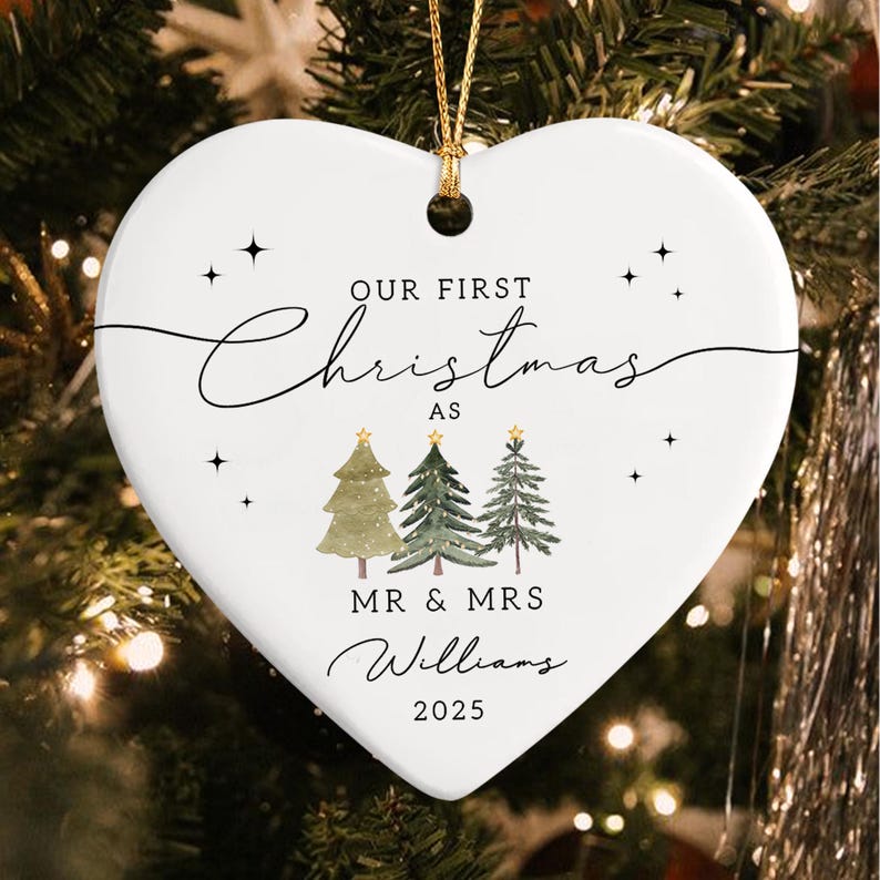 Heart-shaped Christmas ornament with gold string. The white ceramic ornament features the text "Our First Christmas as Mr & Mrs Williams 2025" in black script. Three watercolor Christmas trees are below the text.