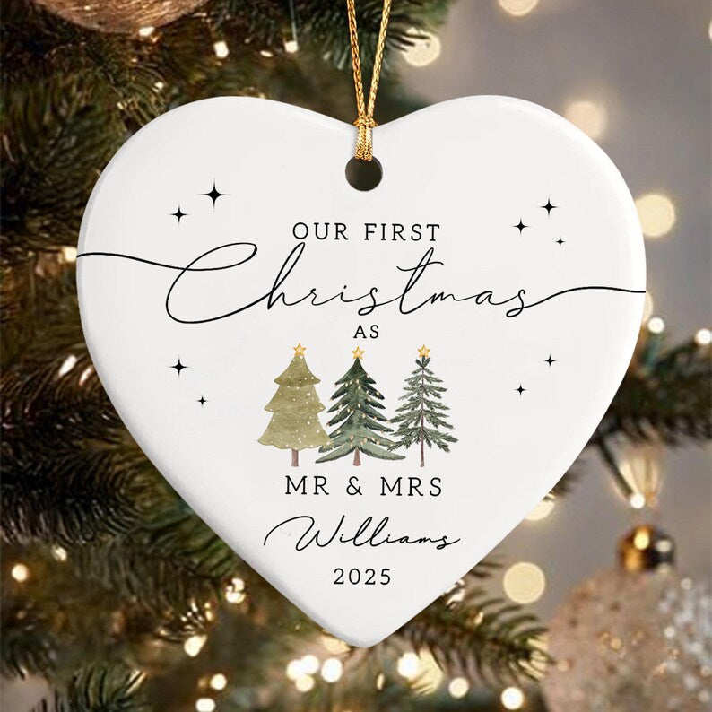 A white heart-shaped Christmas ornament with gold string. The ornament features the text "OUR FIRST Christmas AS MR & MRS Williams 2025" in black script. Three watercolor Christmas trees are below the text.