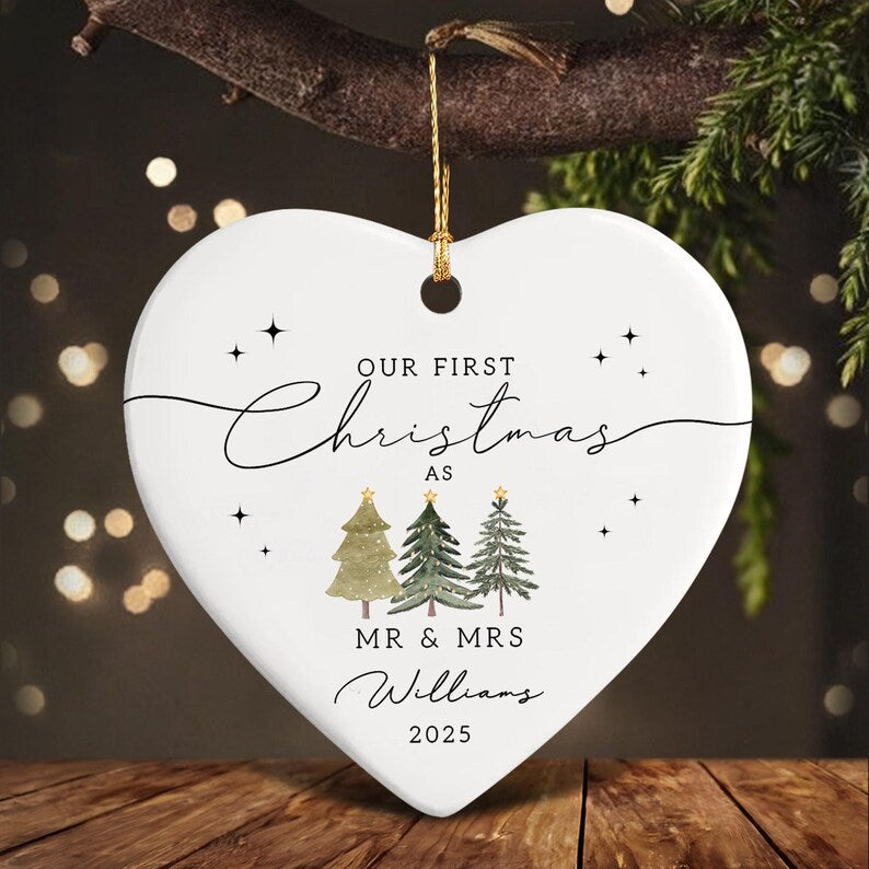 A heart-shaped Christmas ornament with the text "OUR FIRST Christmas AS MR & MRS Williams 2025". The ornament features three watercolor Christmas trees and small star accents. It hangs from a gold string.