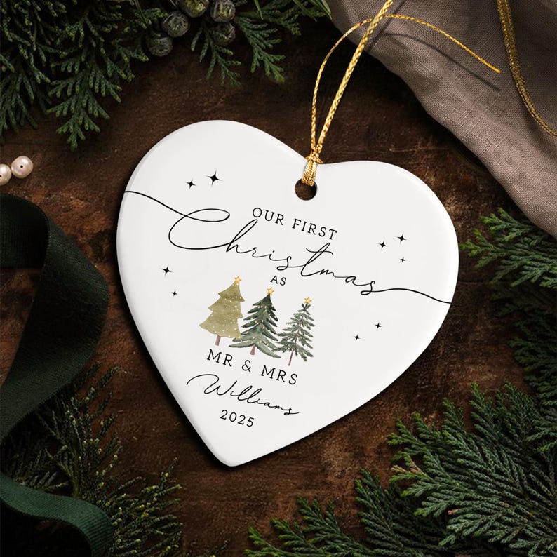 A white heart-shaped Christmas ornament with gold string. The ornament features the text "OUR FIRST Christmas AS MR & MRS Williams 2025" with watercolor Christmas trees and star accents. A festive holiday decoration.