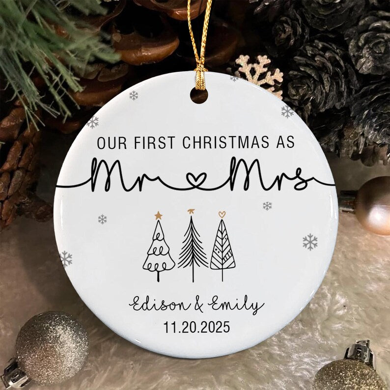 A round white ceramic Christmas ornament featuring the black text "OUR FIRST CHRISTMAS AS Mr & Mrs" with a heart symbol, three stylized black and gold pine trees, and the names "Edison & Emily" with the date "11.20.2025" below.