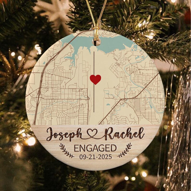 A round ceramic ornament featuring a cream-colored map with a red heart, a vertical line, and a light blue water section. The bottom section has a wood grain texture with the text Joseph & Rachel, ENGAGED, and 09-21-2025.