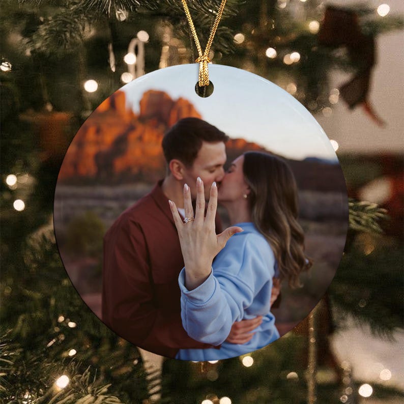 A round ceramic ornament featuring a photograph of a couple embracing outdoors at sunset. The person in the foreground wears a light blue sweatshirt and displays a ring on their finger. A gold string is attached to the top.