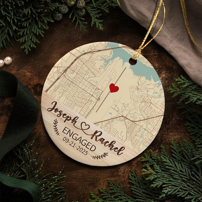 A round ceramic ornament featuring a printed street map with a red heart icon. The lower section has a wood grain pattern with the text Joseph & Rachel, ENGAGED, and the date 09-21-2025, accented by small leaf illustrations.