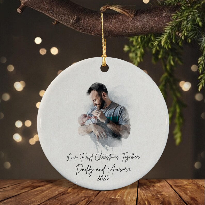 A round white ceramic ornament featuring a watercolor-style image of a person holding a baby. Below the image, the text reads: Our First Christmas Together, Daddy and Aurora, 2025. The ornament hangs from a gold string.