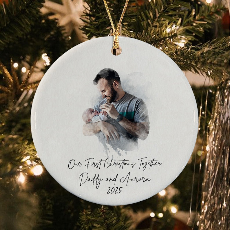 A round white ceramic ornament featuring a watercolor-style illustration of a person holding a baby. Below the image, the text reads: Our First Christmas Together, Daddy and Aurora, 2025. A gold string is attached to the top.
