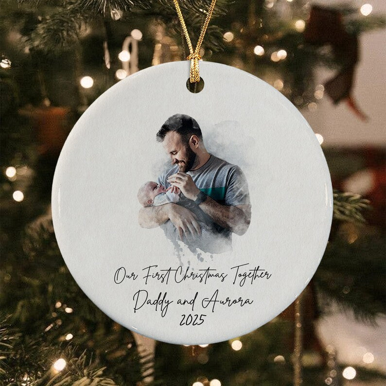 A round white ceramic ornament featuring a watercolor-style illustration of a person holding a baby. The text reads: Our First Christmas Together, Daddy and Aurora, 2025.