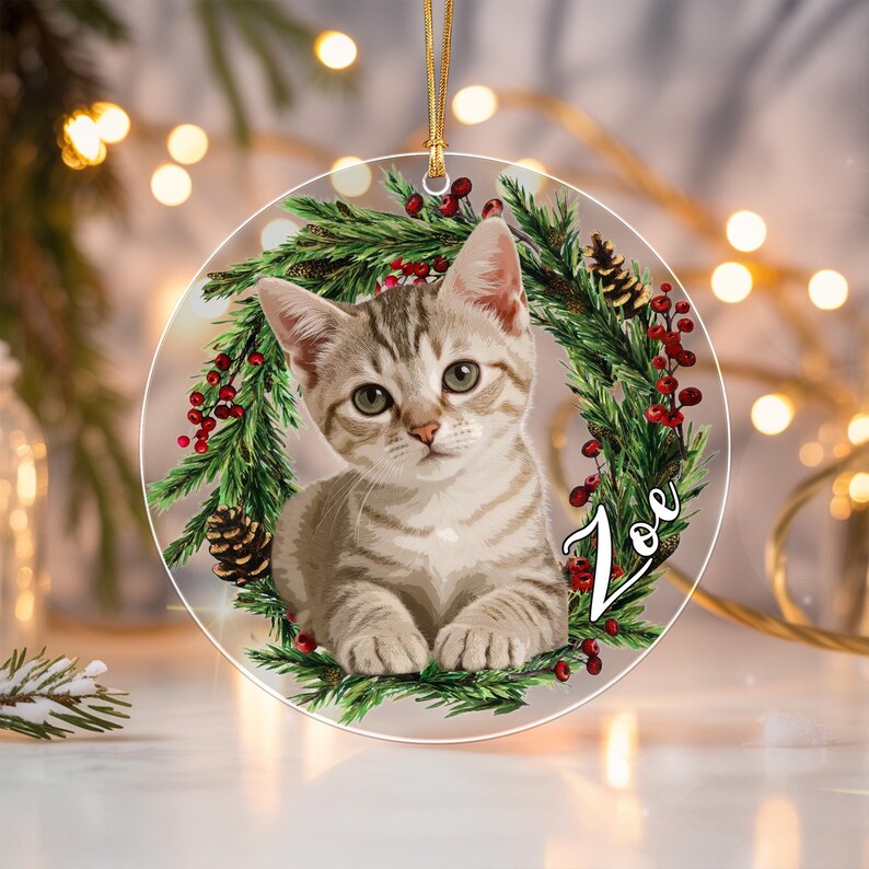 A round, clear acrylic ornament featuring a gray tabby kitten centered within a green pine wreath decorated with red berries and pinecones. The name Zoe is printed in white script on the right side of the wreath.