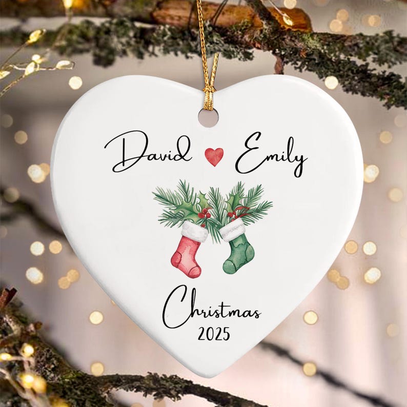 A white heart-shaped ceramic ornament hanging by a gold string. It features the names David and Emily separated by a red heart, a watercolor illustration of two Christmas stockings with pine branches, and the text Christmas 2025.
