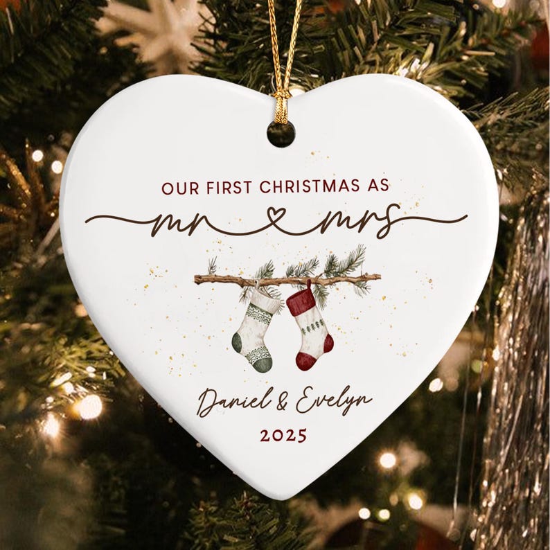 A white heart-shaped ceramic ornament featuring the text "OUR FIRST CHRISTMAS AS mr & mrs" above an illustration of two stockings hanging from a pine branch, with the names "Daniel & Evelyn" and the year "2025" printed below.