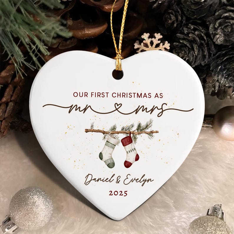 A white heart-shaped ceramic ornament with a gold hanging string. It features the text "OUR FIRST CHRISTMAS AS mr & mrs", "Daniel & Evelyn", and "2025". The design includes a branch with two hanging Christmas stockings.