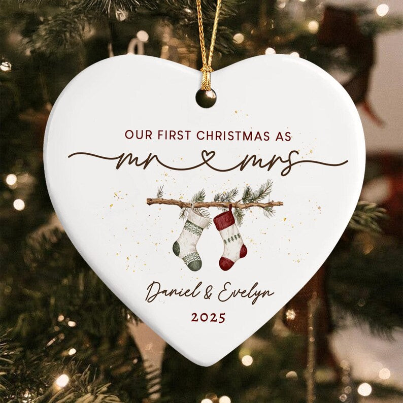 A white heart-shaped ceramic ornament with a gold hanging string. It features the text "OUR FIRST CHRISTMAS AS mr & mrs", "Daniel & Evelyn", and "2025". The design includes a pine branch with two hanging stockings.