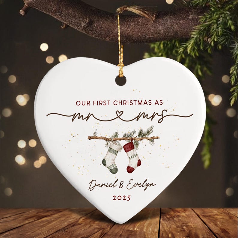 A white heart-shaped ceramic ornament featuring the text "OUR FIRST CHRISTMAS AS mr & mrs", "Daniel & Evelyn", and "2025". The design includes a graphic of two stockings hanging from a pine branch and a gold string.