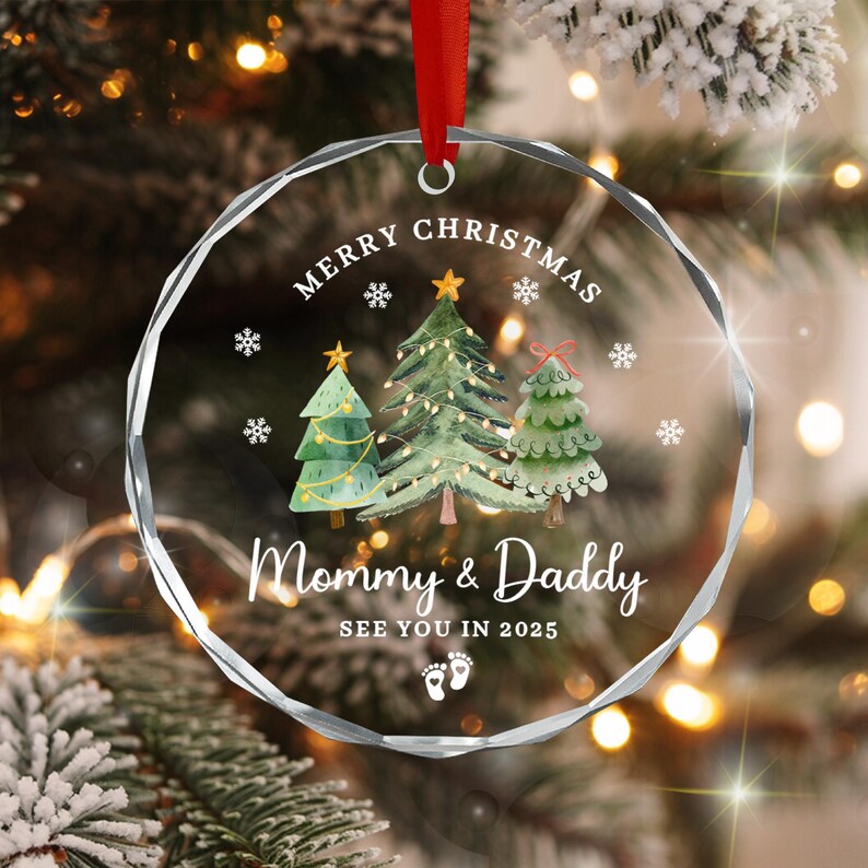 A clear, round Christmas ornament with a red ribbon. The ornament features three illustrated Christmas trees, snowflakes, and the text "MERRY CHRISTMAS" and "Mommy & Daddy SEE YOU IN 2025".