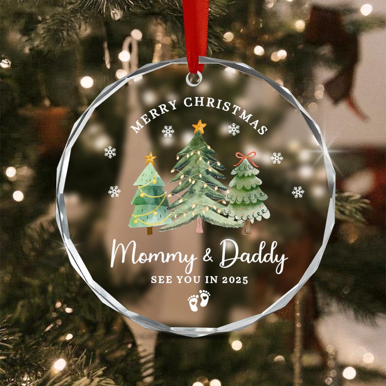 Clear glass Christmas ornament with a red ribbon. The ornament features three illustrated Christmas trees, snowflakes, and the text "Merry Christmas, Mommy & Daddy, See You in 2025" with baby footprints.
