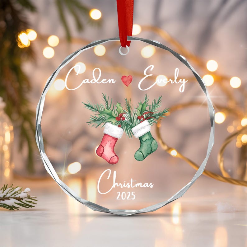 A circular clear acrylic Christmas ornament with a faceted edge, hanging from a red ribbon. It features the names Caden and Everly separated by a red heart, two stockings with holly, and the text Christmas 2025.