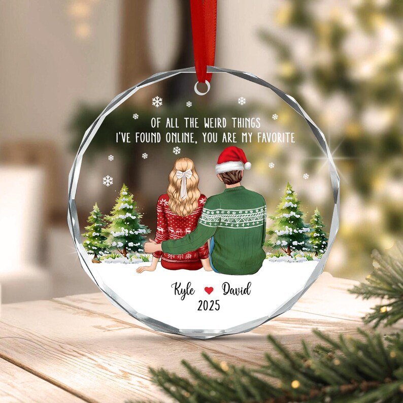 A round, clear acrylic Christmas ornament featuring a couple sitting in snow with pine trees. The text reads: OF ALL THE WEIRD THINGS I'VE FOUND ONLINE, YOU ARE MY FAVORITE. Below are the names Kyle and David with 2025.