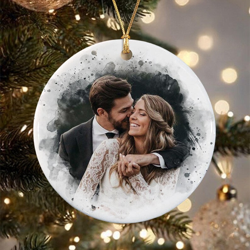 A round white ceramic ornament featuring a watercolor-style portrait of a couple embracing. The person on the left wears a dark suit, while the person on the right wears a white lace dress. A gold string is attached to the top.