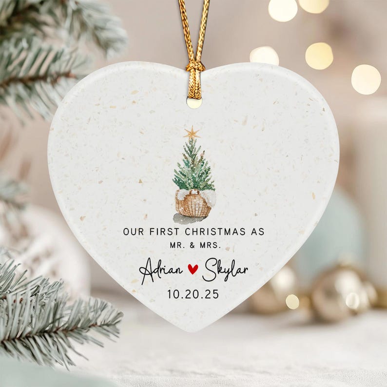 A white heart-shaped ceramic ornament with a gold string, featuring a watercolor illustration of a small Christmas tree in a basket, the text "OUR FIRST CHRISTMAS AS MR. & MRS.", the names "Adrian" and "Skylar" with a red heart.
