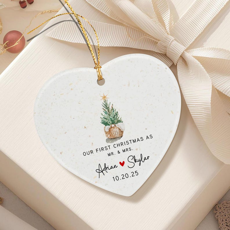 A heart-shaped white ceramic ornament with a speckled texture, featuring a watercolor illustration of a Christmas tree in a basket, the text "OUR FIRST CHRISTMAS AS MR. & MRS.", the names "Adrian" and "Skylar" with a red heart.