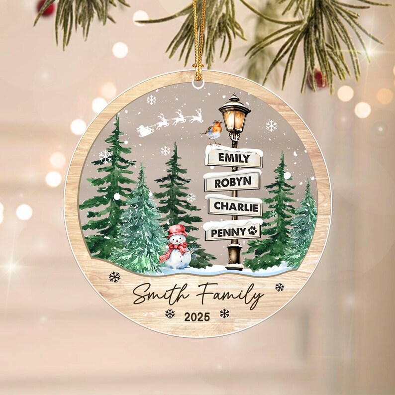 A round Christmas ornament featuring a winter scene with green pine trees, a snowman, and a street lamp with signs reading EMILY, ROBYN, CHARLIE, and PENNY. The text Smith Family and 2025 are printed below.