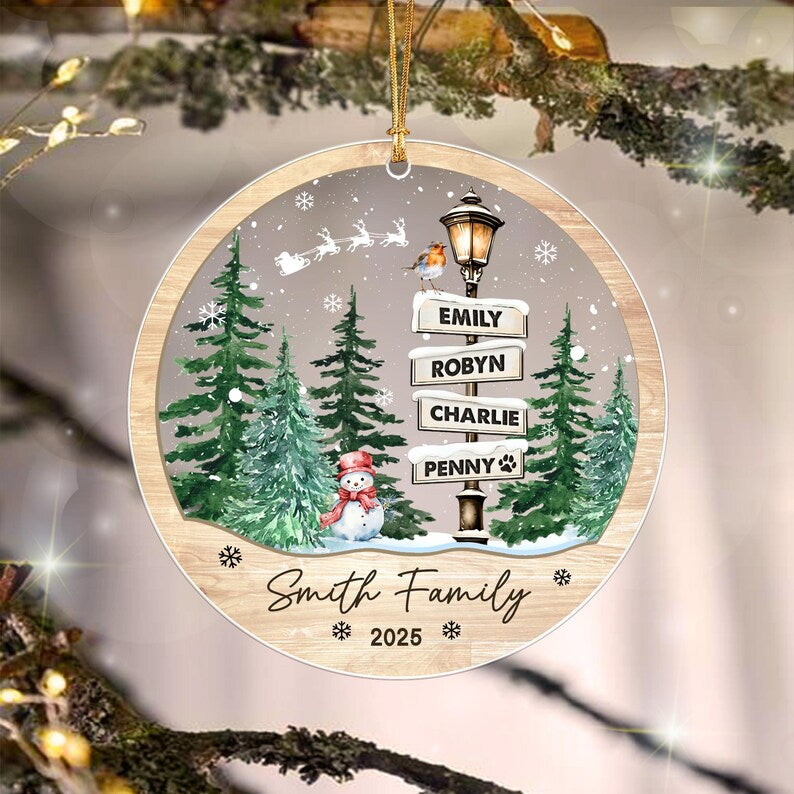 A round Christmas ornament featuring a winter scene with green pine trees, a snowman, and a street lamp with four signs labeled Emily, Robyn, Charlie, and Penny.
