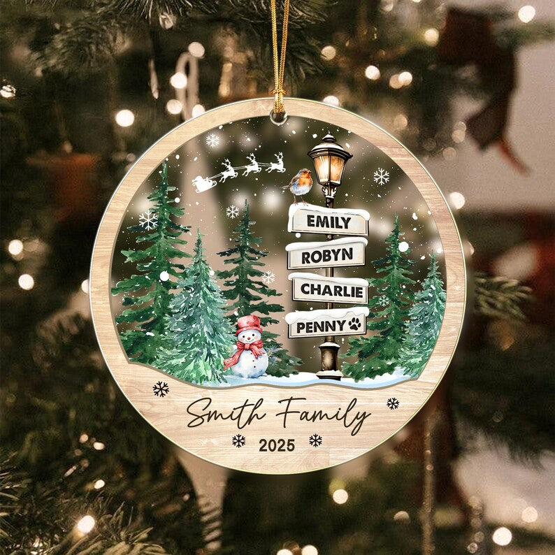 A round, wood-textured Christmas ornament featuring a winter scene with green pine trees, a snowman, a street lamp with signs reading EMILY, ROBYN, CHARLIE, and PENNY, a robin, a flying sleigh, and the text Smith Family 2025.
