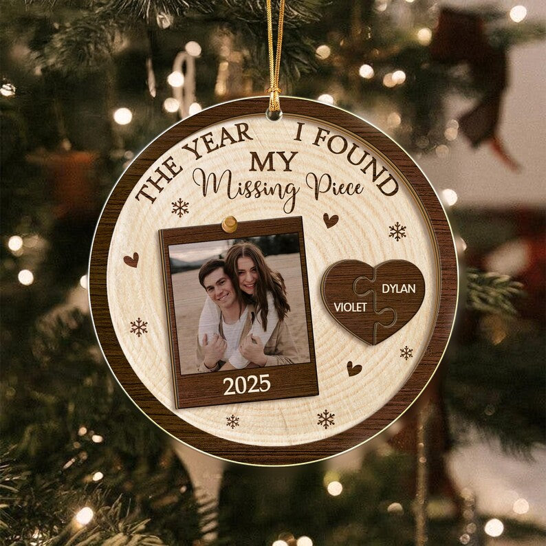 A round wooden Christmas ornament featuring the text THE YEAR I FOUND MY Missing Piece and 2025. It displays a square photo of two people and a dark brown heart-shaped puzzle piece with the names VIOLET and DYLAN engraved.