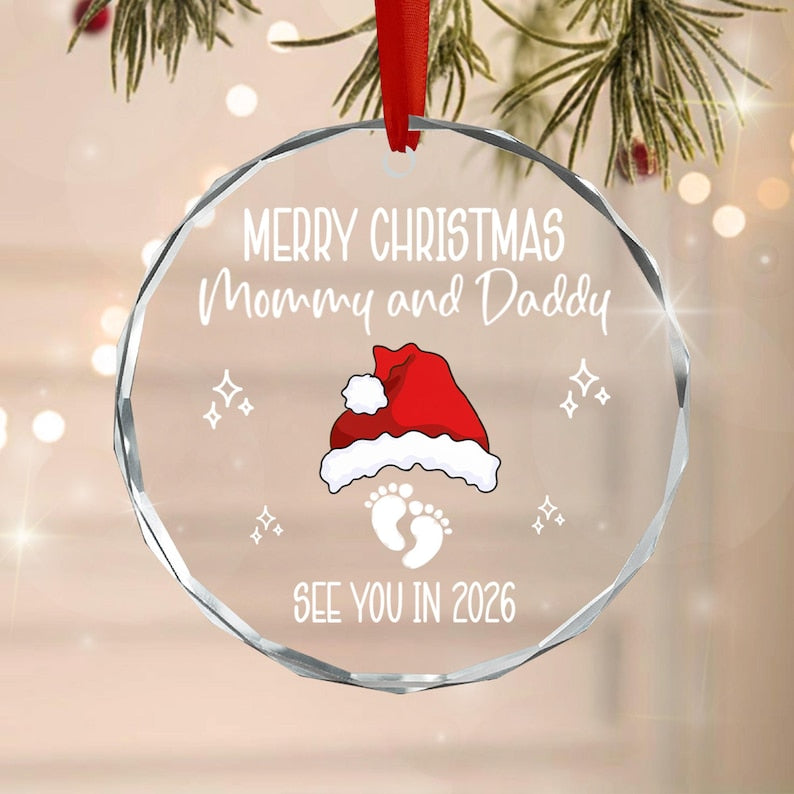 A round, clear, faceted glass Christmas ornament hanging from a red ribbon. The text reads Merry Christmas Mommy and Daddy, See You In 2026. A red and white Santa hat graphic sits above two white baby footprints and four stars.