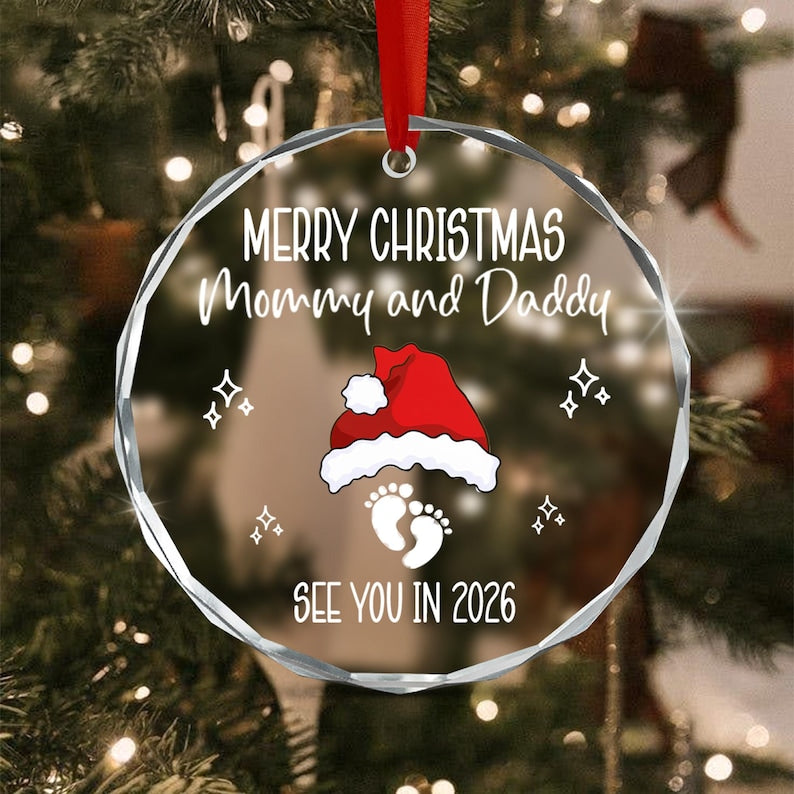 A round, clear, faceted glass Christmas ornament hanging from a red ribbon. It features white text reading Merry Christmas Mommy and Daddy, a red and white Santa hat, white baby footprints, and the text See You In 2026.