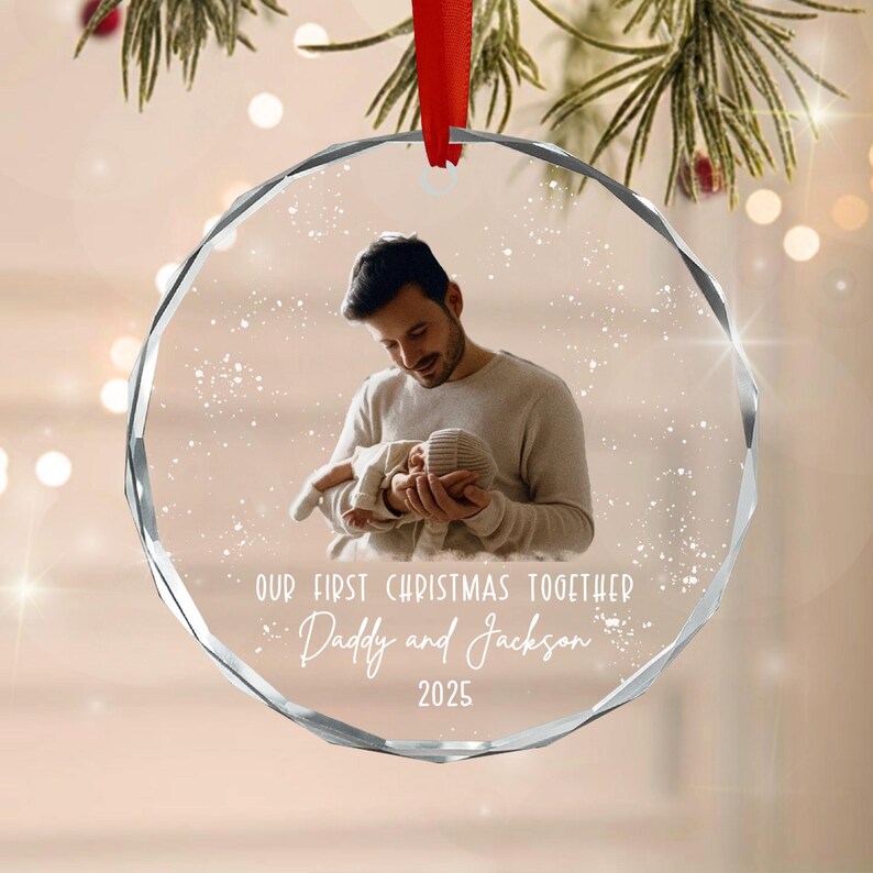 A round, faceted glass Christmas ornament featuring a printed photograph of a person holding a baby. The text reads: OUR FIRST CHRISTMAS TOGETHER, Daddy and Jackson, 2025. A red ribbon is attached to the top for hanging.