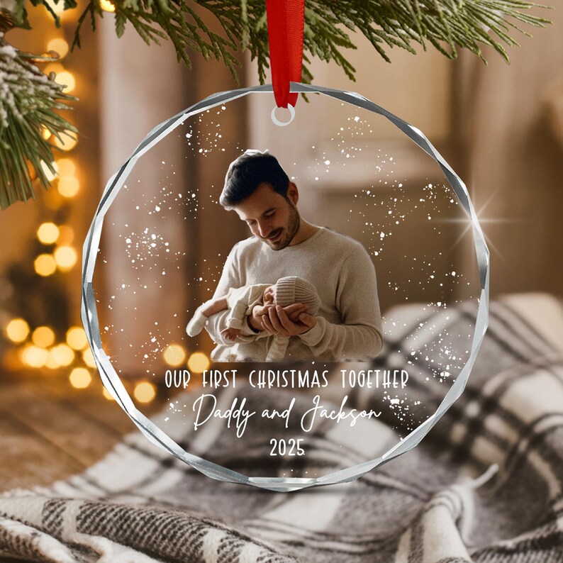 A circular clear acrylic ornament with faceted edges, featuring a printed photograph of a person holding a baby. The text reads: OUR FIRST CHRISTMAS TOGETHER, Daddy and Jackson, 2025. A red ribbon is attached to the top.