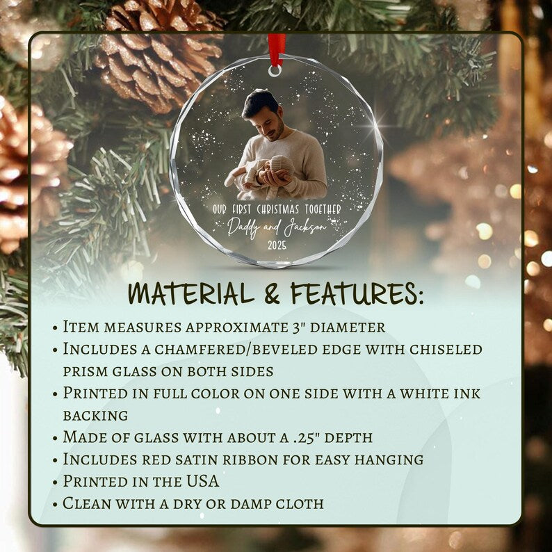 A circular glass Christmas ornament with a beveled edge, featuring a printed photograph of a person holding an infant. The text reads Our First Christmas, Daddy and Jackson, 2025. It includes a red satin ribbon for hanging.