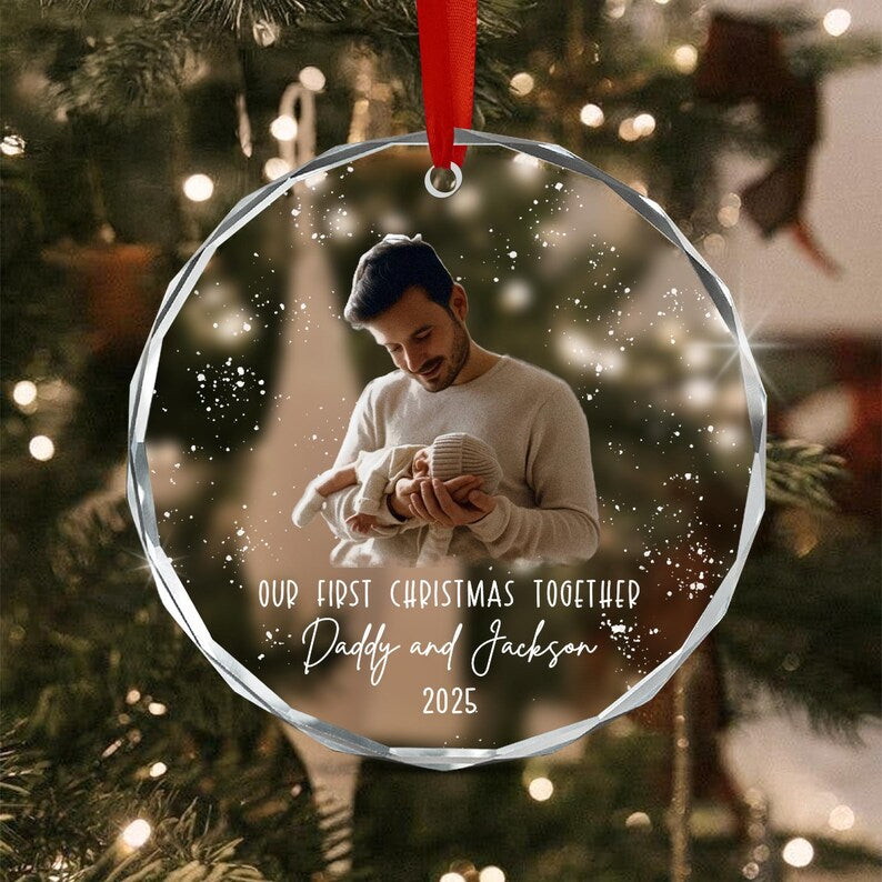 A round, clear, faceted glass ornament featuring a printed photograph of a person holding a baby. The text reads: OUR FIRST CHRISTMAS TOGETHER, Daddy and Jackson, 2025. A red ribbon is attached to the top for hanging.