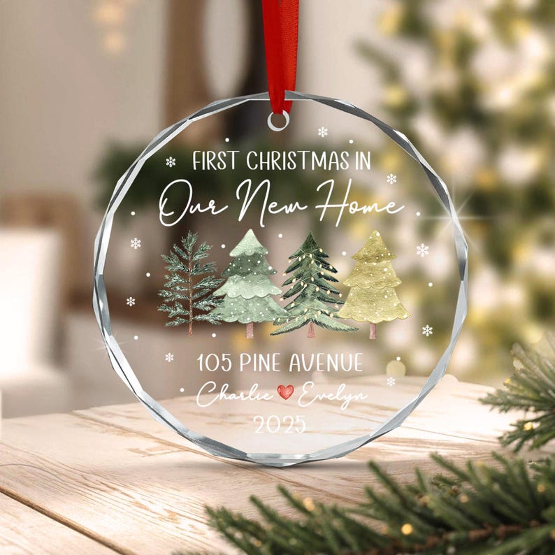 A circular clear glass Christmas ornament with a red ribbon hanger. The design features four watercolor-style pine trees, white snowflakes, and the text: FIRST CHRISTMAS IN Our New Home, 105 PINE AVENUE, Charlie, Evelyn, and 2025.