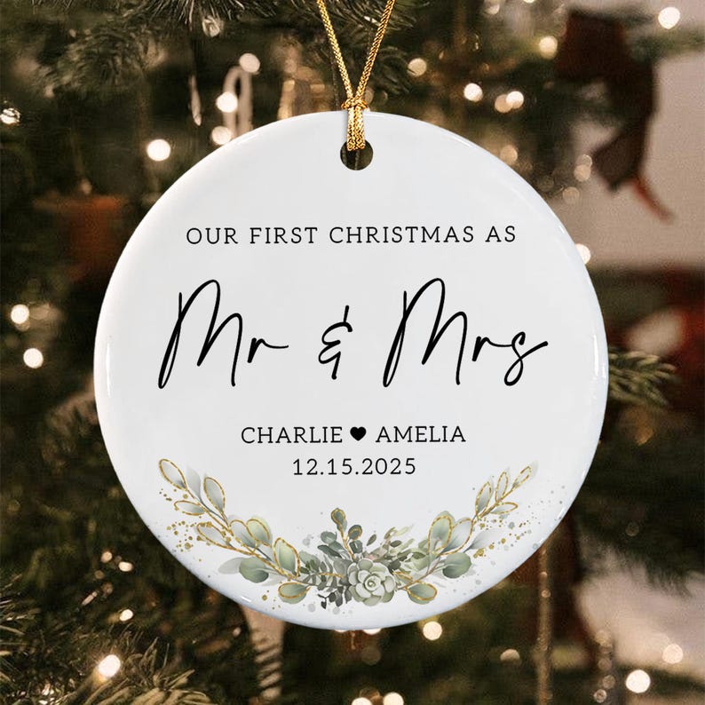 A round white ceramic Christmas ornament featuring the black text "OUR FIRST CHRISTMAS AS Mr & Mrs" followed by the names "CHARLIE ♥ AMELIA" and the date "12.15.2025" above a decorative illustration of green leaves and succulents.