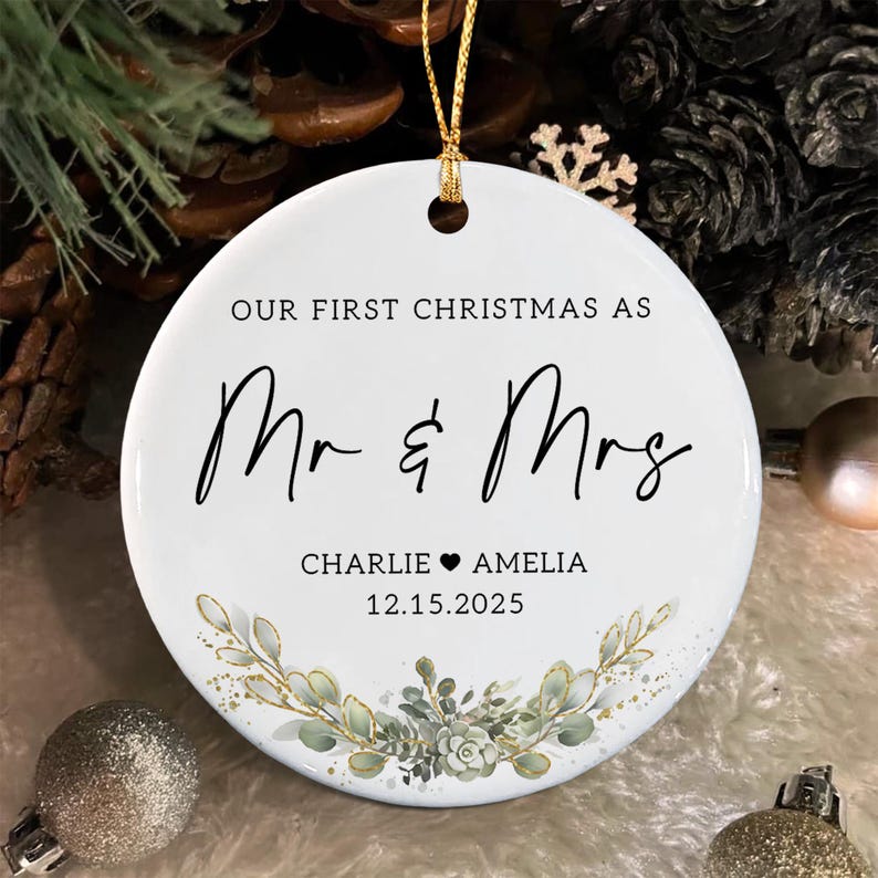 A round white ceramic ornament featuring the black text OUR FIRST CHRISTMAS AS Mr & Mrs, followed by the names CHARLIE and AMELIA separated by a heart, and the date 12.15.2025.