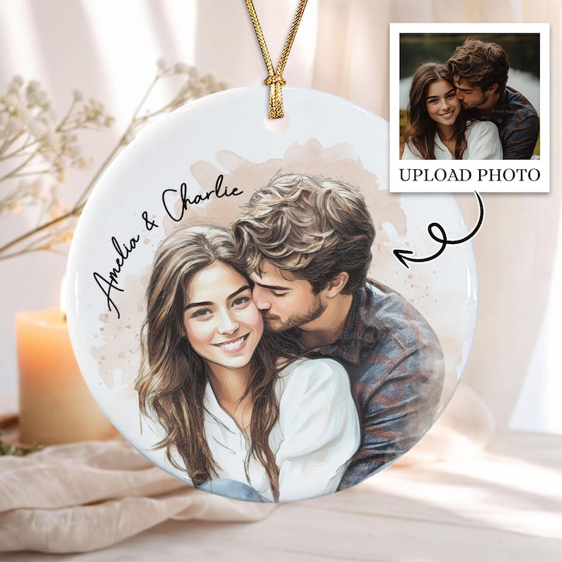 A round white ceramic ornament featuring a custom watercolor-style illustration of a couple, with the names Amelia & Charlie printed in black script. The ornament hangs from a gold cord and includes a photo upload placeholder.