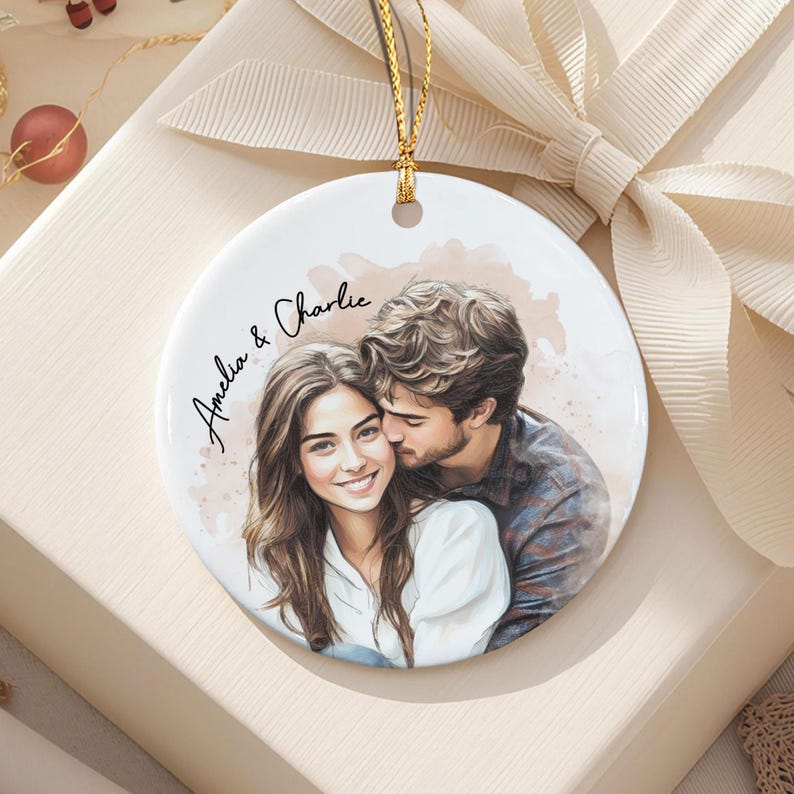 A round white ceramic ornament featuring a watercolor-style illustration of a couple embracing. The text "Amelia & Charlie" is printed in black script on the left side. A gold string is attached to the top for hanging purposes.