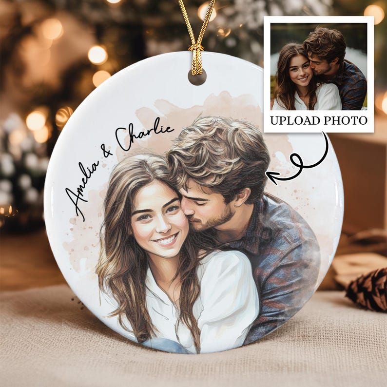 A round white ceramic ornament featuring a custom watercolor-style illustration of a couple. The text "Amelia & Charlie" is printed in black script. A gold cord is attached at the top.
